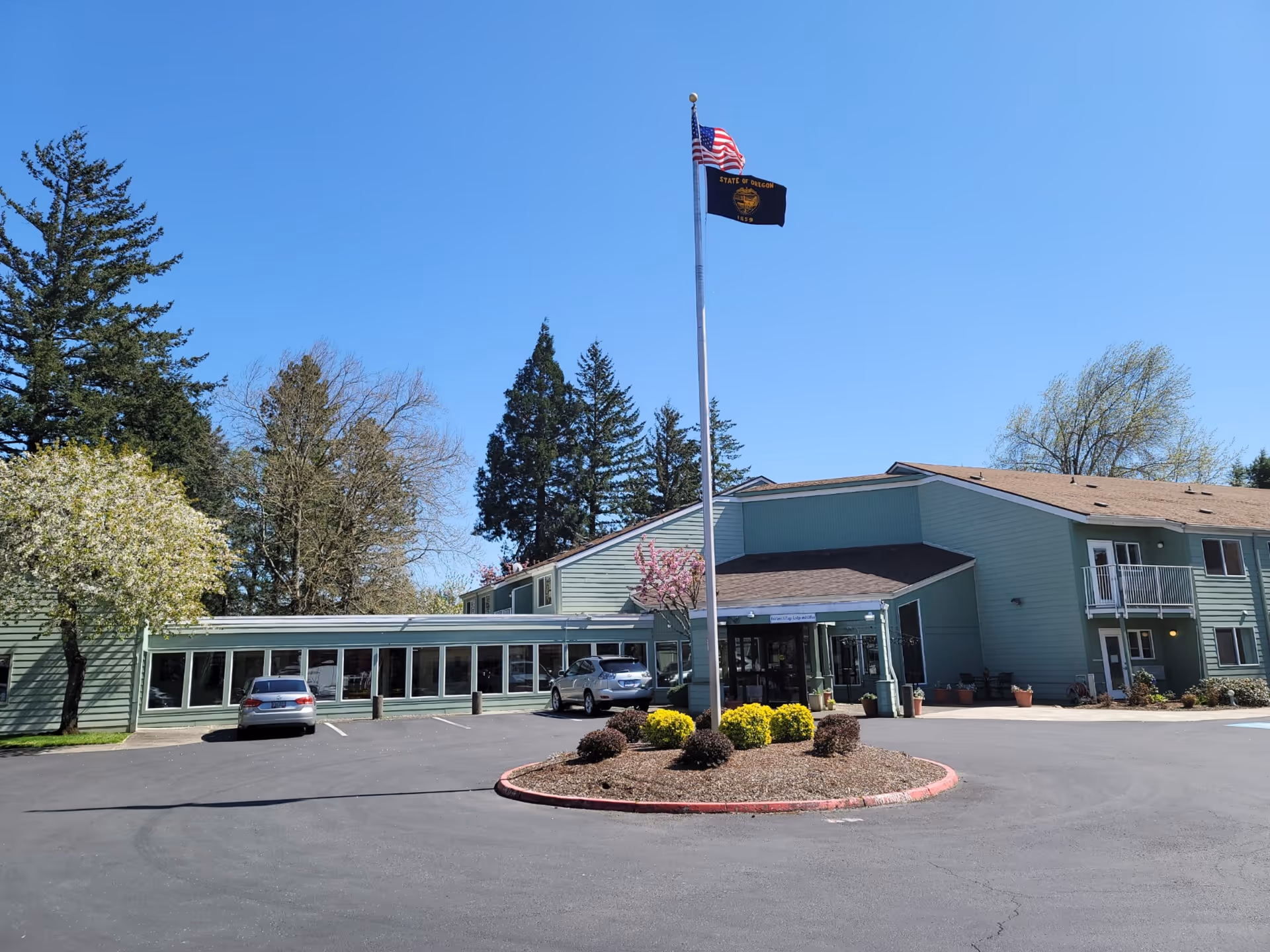 Exterior view of Fairlawn of Olympus Retirement Living facility showing a two-story green building with a parking lot in front, two cars parked, a flagpole with the American flag and the State of Oregon flag, surrounded by trees and clear blue sky.