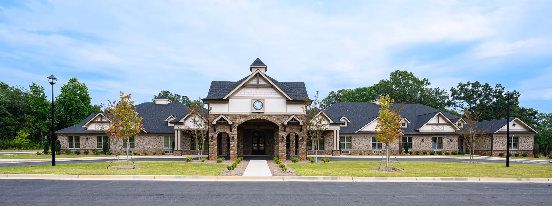 Front exterior of a single-story brick senior living building with a central covered entrance topped by a clock and landscaped lawn.