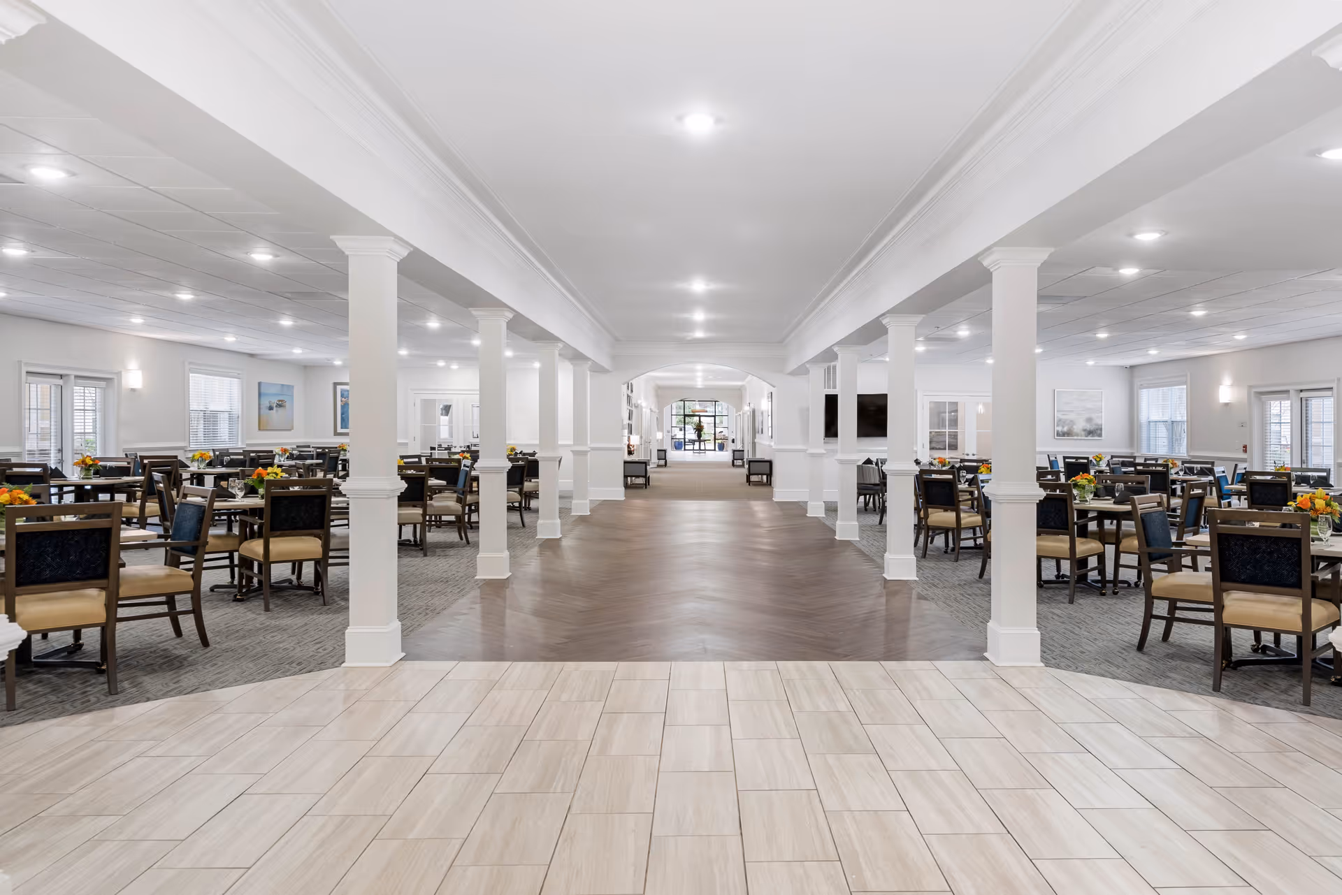 Spacious dining room in a senior living facility with multiple tables and chairs arranged neatly. The room features white walls, columns, and a mix of tile and wood flooring. Each table has a small floral centerpiece, and the area is well-lit with ceiling lights and natural light from windows.