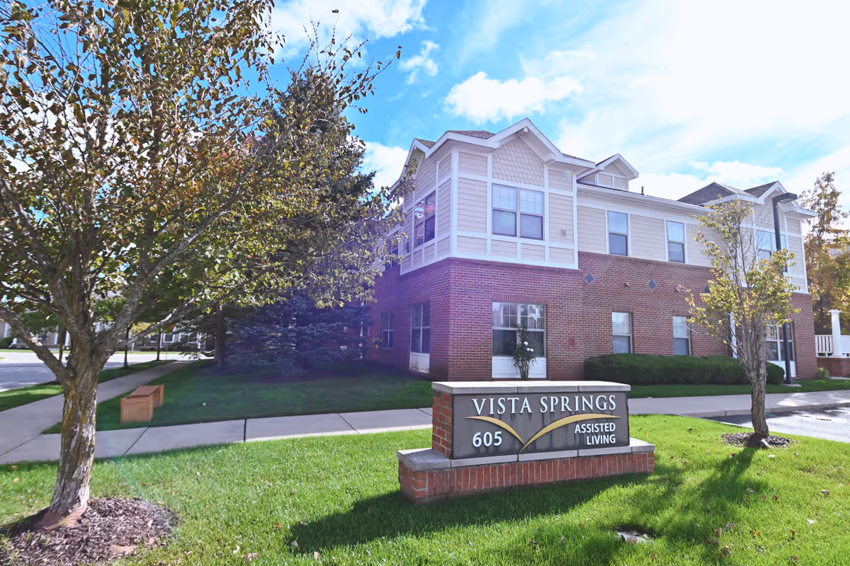 Exterior view of a two-story assisted living building with a brick and beige siding facade, surrounded by green grass and trees under a partly cloudy blue sky. A sign in front reads 'Vista Springs 605 Assisted Living'.