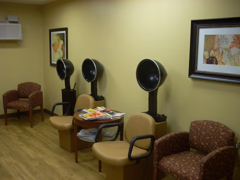 Interior of a salon area in a senior living facility with three vintage hair dryers mounted on stands, two tan salon chairs with armrests, a round wooden table with magazines on it, and two patterned armchairs against yellow walls with framed artwork.