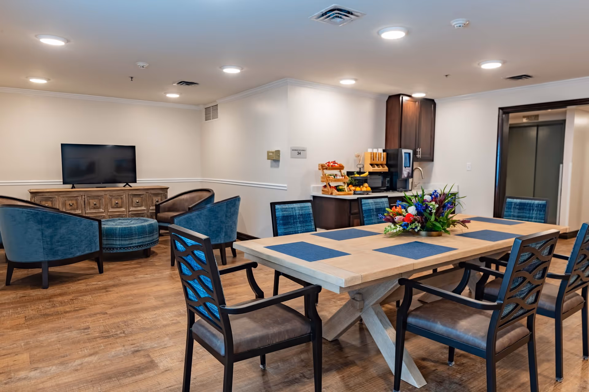 A senior living facility common area featuring a wooden dining table with six chairs, each with blue cushions and placemats. A colorful flower arrangement is centered on the table. In the background, there is a small seating area with four blue upholstered chairs and an ottoman facing a flat-screen TV on a wooden cabinet. To the right, a kitchenette area with dark cabinets, a coffee machine, and a fruit stand is visible. The room has wood flooring, white walls, and recessed ceiling lights.