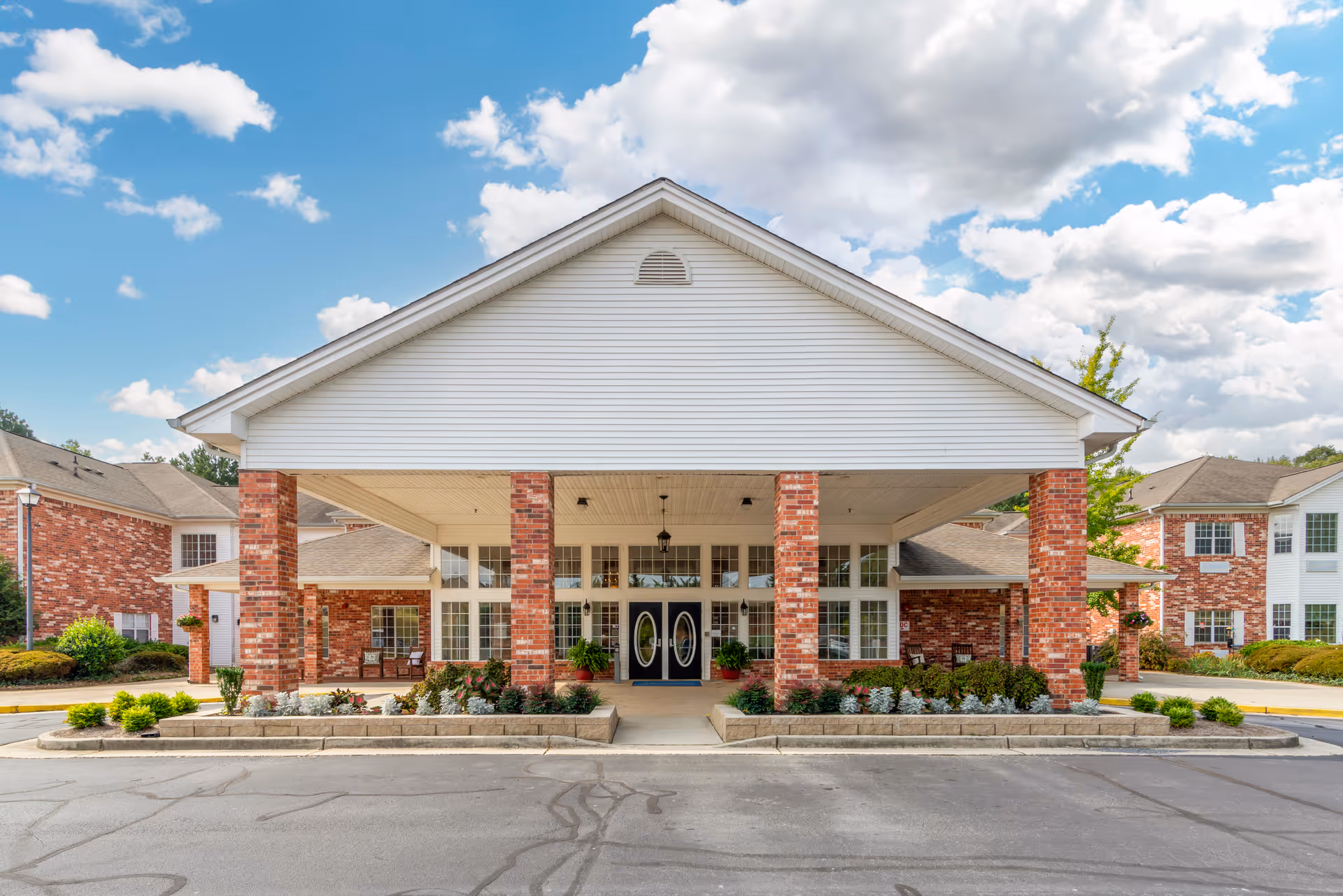 Front exterior view of a senior living facility building with a large covered entrance supported by brick columns, surrounded by landscaped bushes and plants under a partly cloudy sky.
