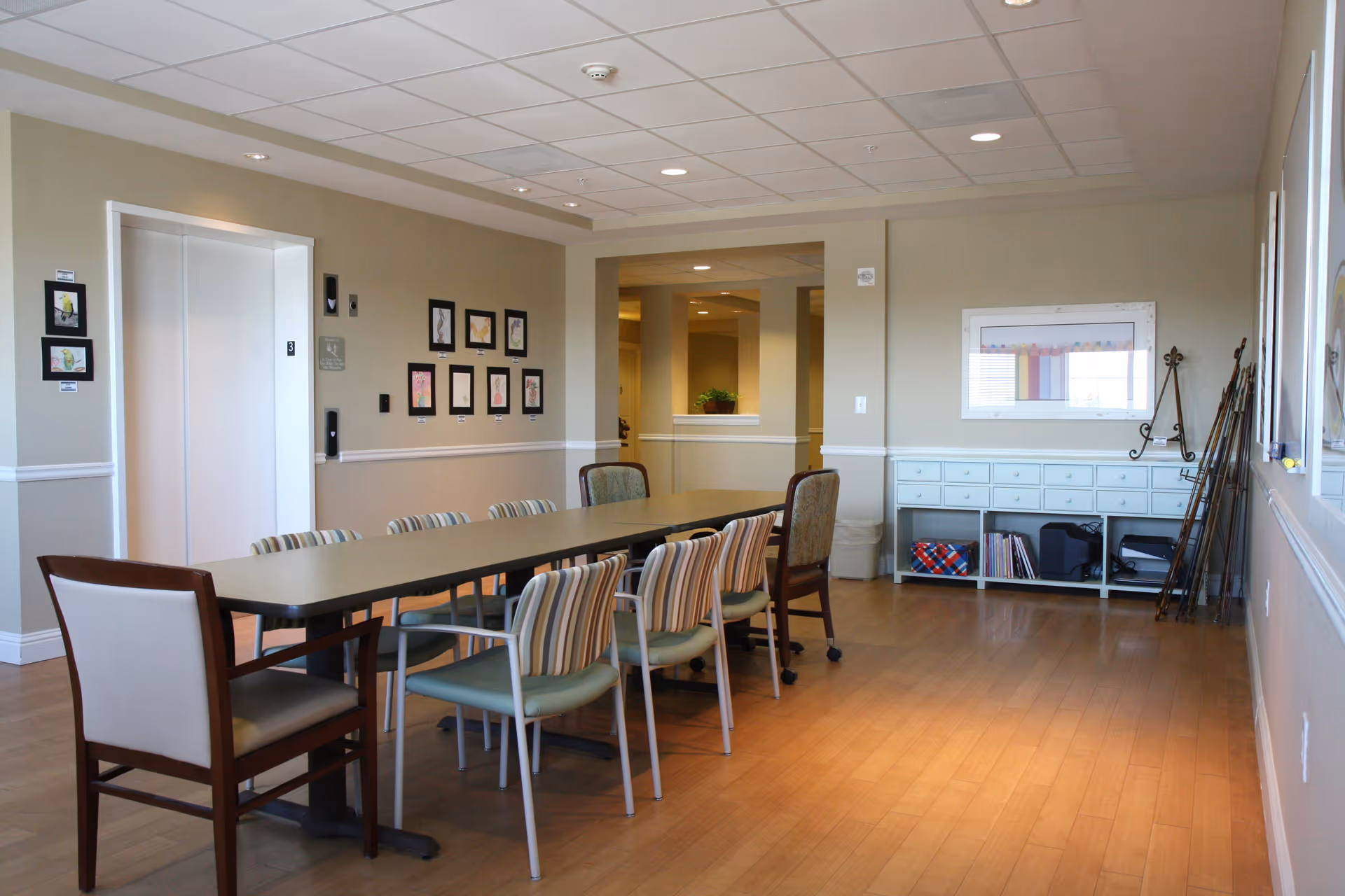 A well-lit room with a long rectangular table surrounded by various chairs, including striped and solid-colored ones. The room has light-colored walls with framed artwork, a white cabinet with multiple drawers, and a window. The floor is wooden, and there is an elevator door on the left side of the room.