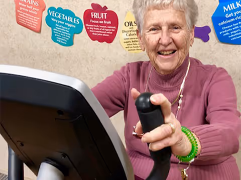 An elderly woman with short white hair wearing a pink sweater is smiling while using an exercise bike. Behind her, colorful posters on the wall display nutritional information about grains, vegetables, fruit, oils, and milk.