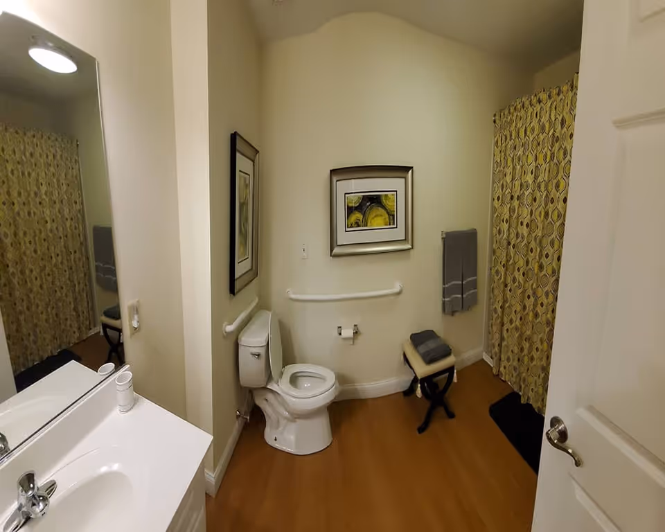A clean and well-lit bathroom with a white toilet, a white sink with a faucet, a large mirror, and a shower area with a yellow patterned curtain. There is a small cushioned stool with folded towels on it, a towel rack with a gray towel, and a framed abstract artwork on the wall. The floor is wooden, and the walls are painted a light beige color.