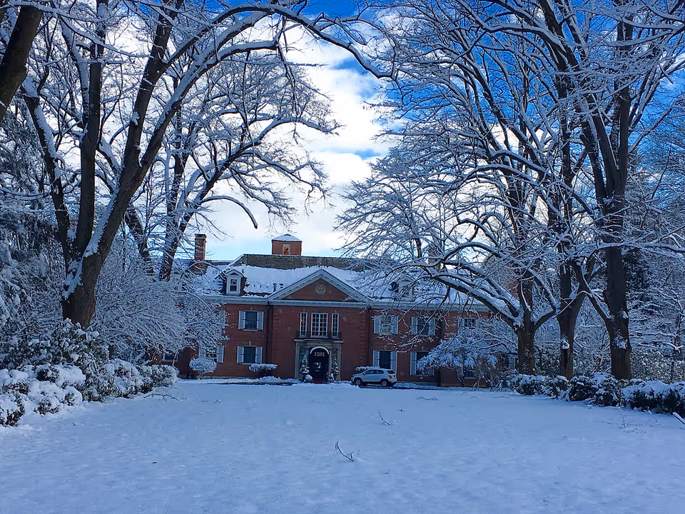 A large brick building with a snow-covered roof and surrounding trees also covered in snow. The ground is blanketed with fresh snow, and a car is parked near the entrance of the building under a partly cloudy blue sky.