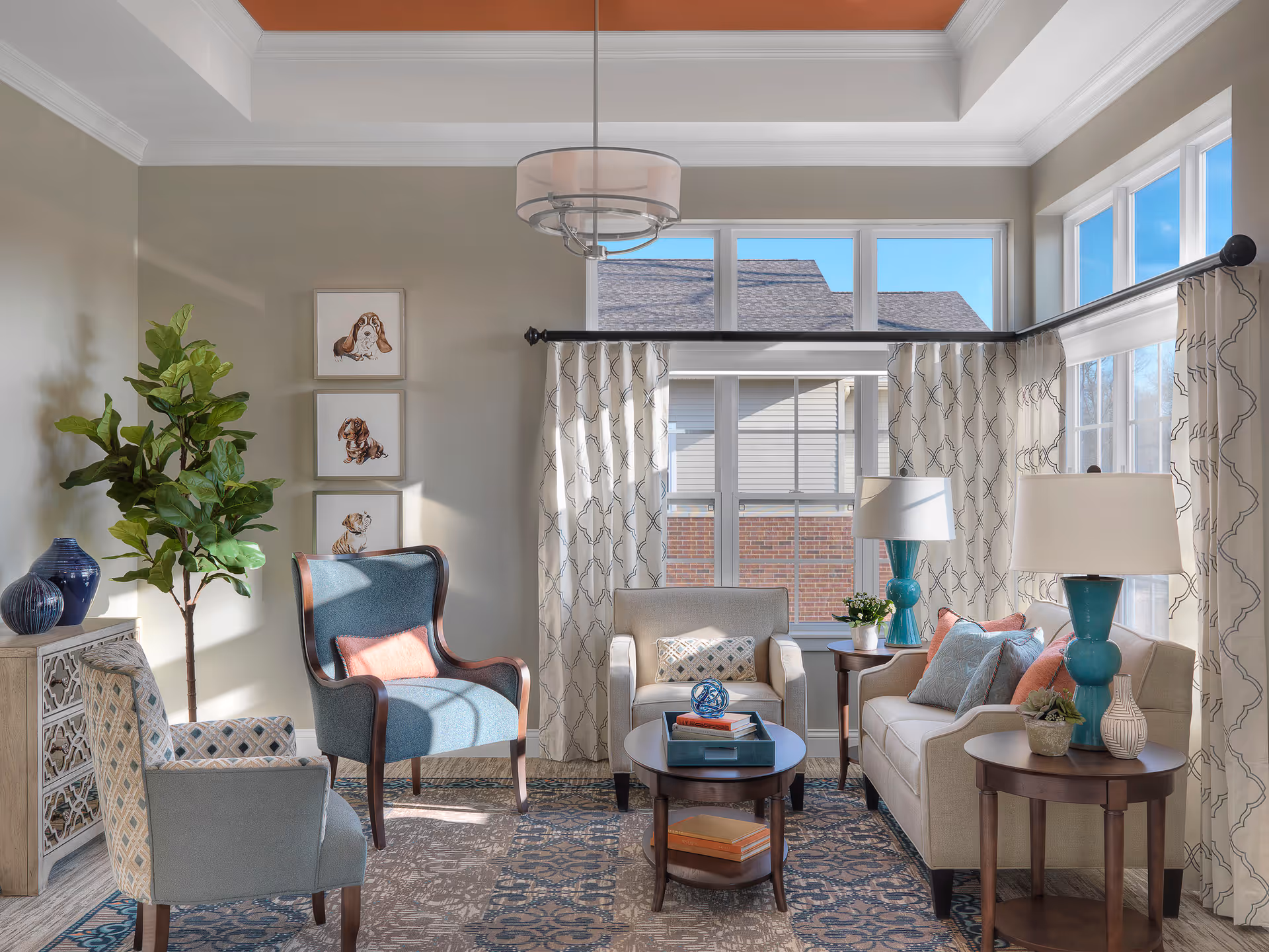 A bright and cozy living room with large windows letting in natural light. The room features a beige sofa with colorful cushions, two armchairs, a round coffee table with books and decorative items, two side tables with teal lamps, a patterned rug, and a tall green plant in the corner. Three framed dog illustrations hang on the wall.