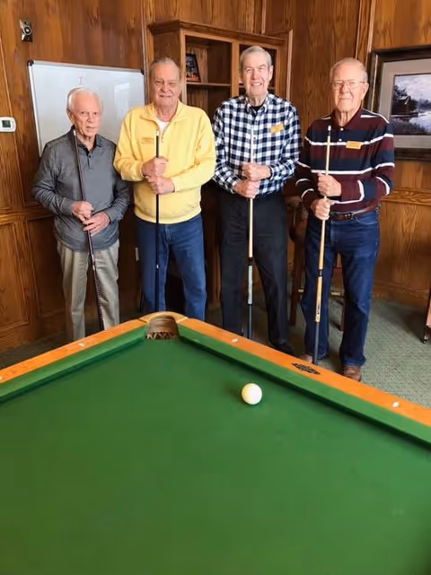 Four elderly men standing behind a pool table in a wood-paneled room, each holding a pool cue. The pool table has a green felt surface with a single white cue ball near the corner pocket.