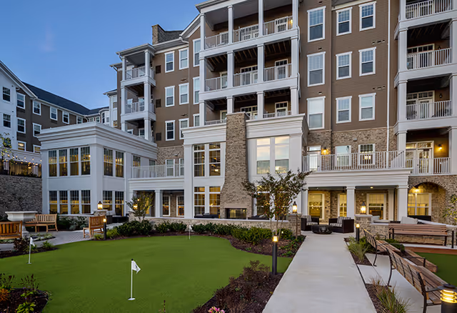Exterior view of a multi-story senior living facility with balconies and large windows. In the foreground, there is a well-maintained putting green surrounded by benches, plants, and a paved walkway leading to the building entrance. Outdoor seating areas with chairs and tables are visible near the building.
