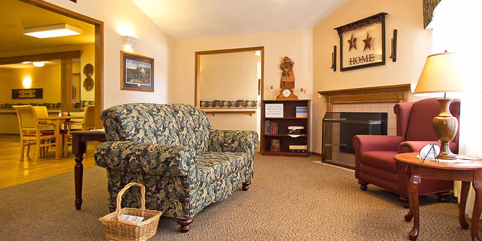 Comfortable living room with a floral loveseat, a red armchair by a fireplace, a side table with a lamp, and a dining area visible through a doorway.