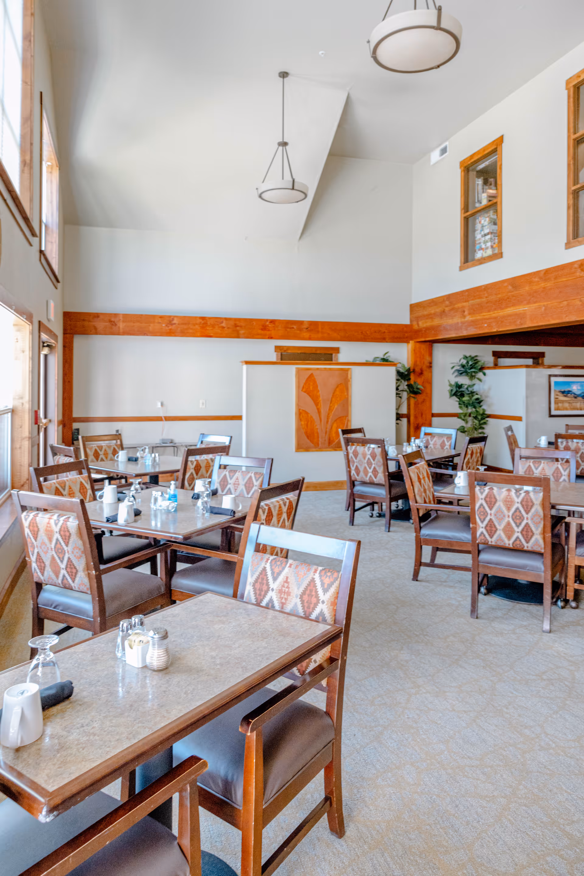 A bright dining room with multiple tables and chairs arranged neatly. The room features high ceilings with two hanging light fixtures, large windows on the left side allowing natural light to fill the space, and wooden accents along the walls and furniture. Tables are set with salt and pepper shakers, napkins, and glasses. There are some plants and framed pictures on the walls in the background.