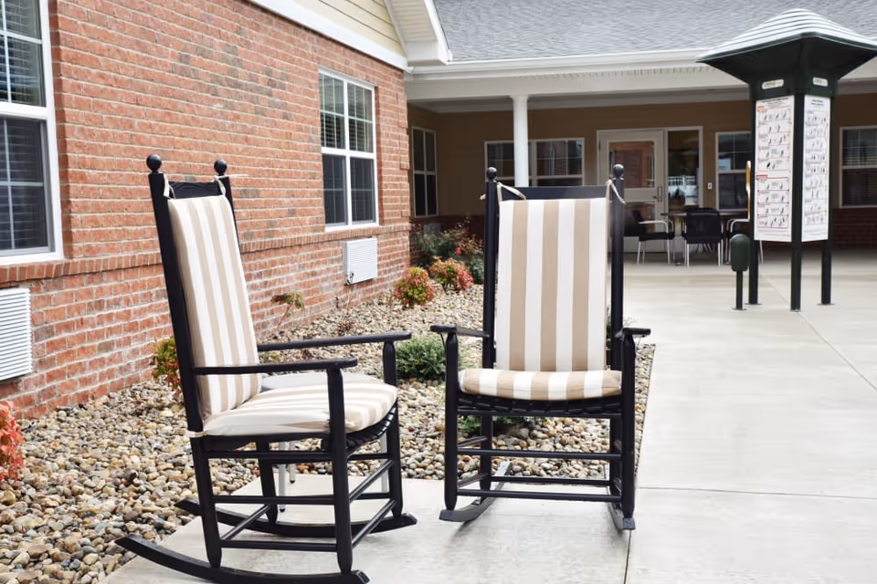 Two black wooden rocking chairs with beige and white striped cushions placed on a concrete patio outside a brick building with windows. There are small plants and rocks along the building's foundation, and a covered porch area with additional seating and an exercise chart in the background.
