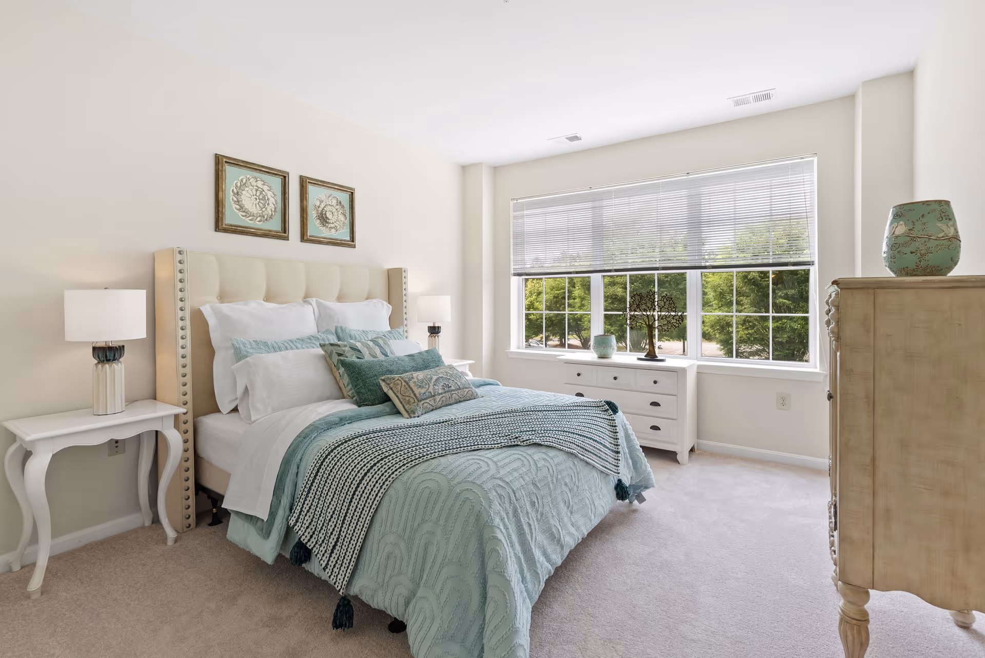 A bright and airy bedroom with a large window letting in natural light. The room features a neatly made bed with multiple pillows and a light blue patterned bedspread. There are two white nightstands with lamps on either side of the bed, two framed artworks above the headboard, a white dresser under the window, and a beige chest of drawers with a decorative vase on top.