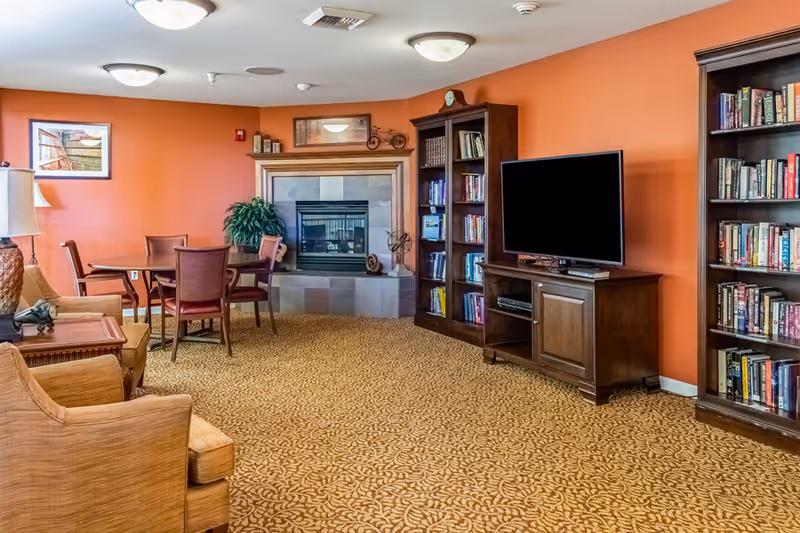 A cozy living room with orange walls, patterned carpet, a fireplace with a wooden mantel, two wooden bookshelves filled with books, a flat-screen TV on a wooden stand, a small round table with four chairs, and comfortable armchairs with a side table and lamp.