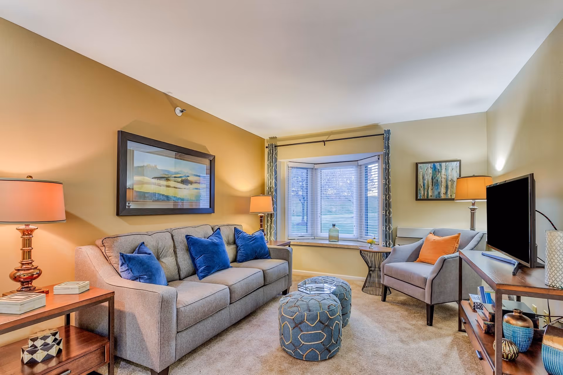 A cozy living room with a beige sofa adorned with blue pillows, a matching armchair with an orange pillow, two blue patterned ottomans, a wooden side table with a lamp, a wooden TV stand with a flat-screen TV, and a large window with blinds and curtains letting in natural light.