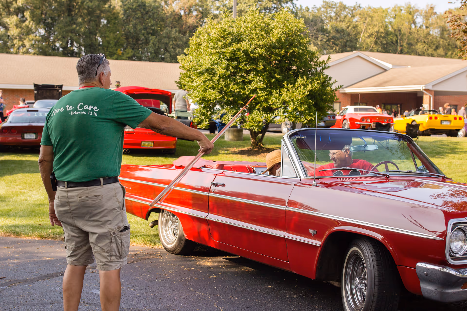 A man wearing a green shirt and beige shorts is standing next to a red classic convertible car, holding a long stick. Another person is sitting inside the car, wearing a hat. In the background, there are more classic cars parked on a grassy area near a building with people gathered around.