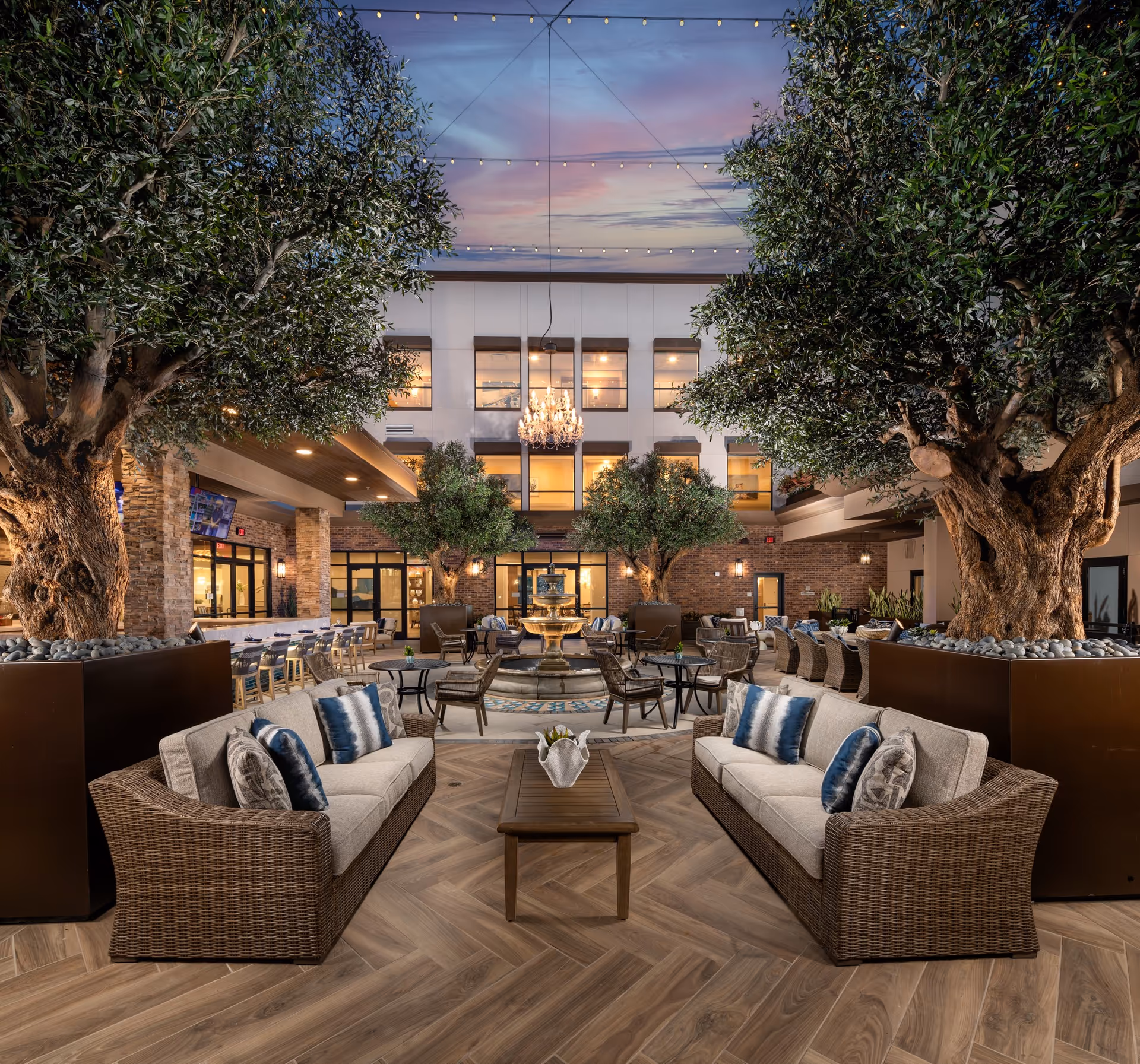 Spacious and elegant outdoor courtyard at Clearwater Newport Beach featuring comfortable wicker sofas with cushions and pillows, large potted trees, a central water fountain, string lights overhead, and a chandelier hanging in the center. The area is surrounded by a building with large windows and warm lighting.