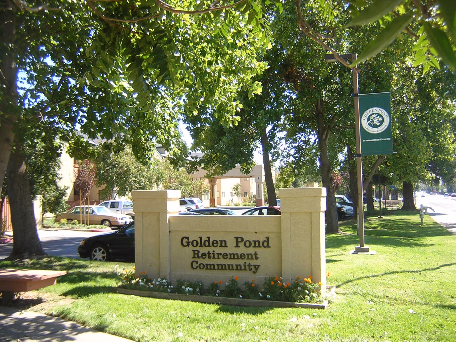 Entrance sign for Golden Pond Retirement Community on a grassy lawn with trees, parked cars, and a lamppost banner.