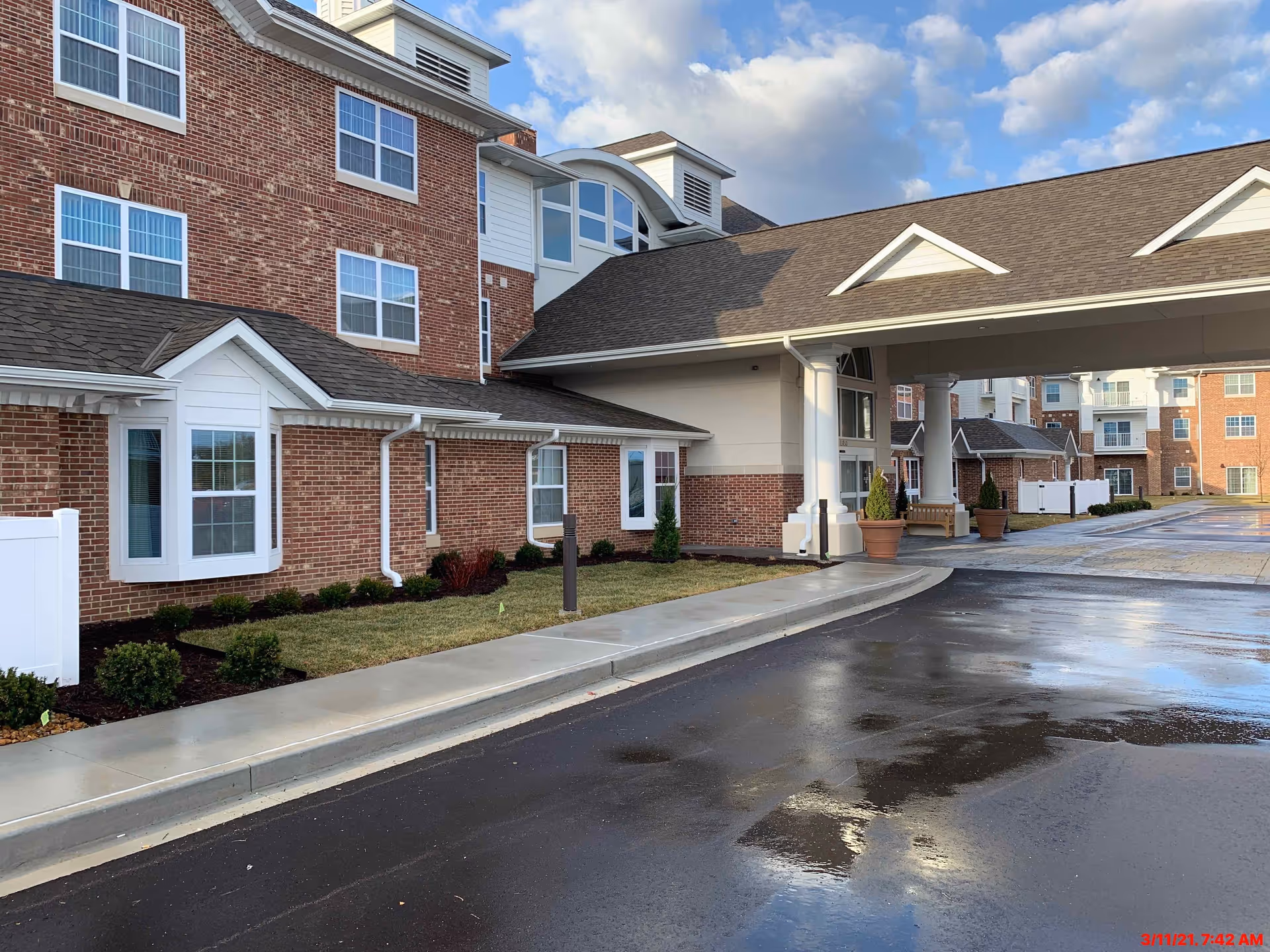 Exterior view of a multi-story brick building with white trim, featuring large windows and a covered entrance with columns. The driveway and sidewalk are wet, reflecting the cloudy sky above. There are potted plants near the entrance and small landscaped areas with shrubs along the building.