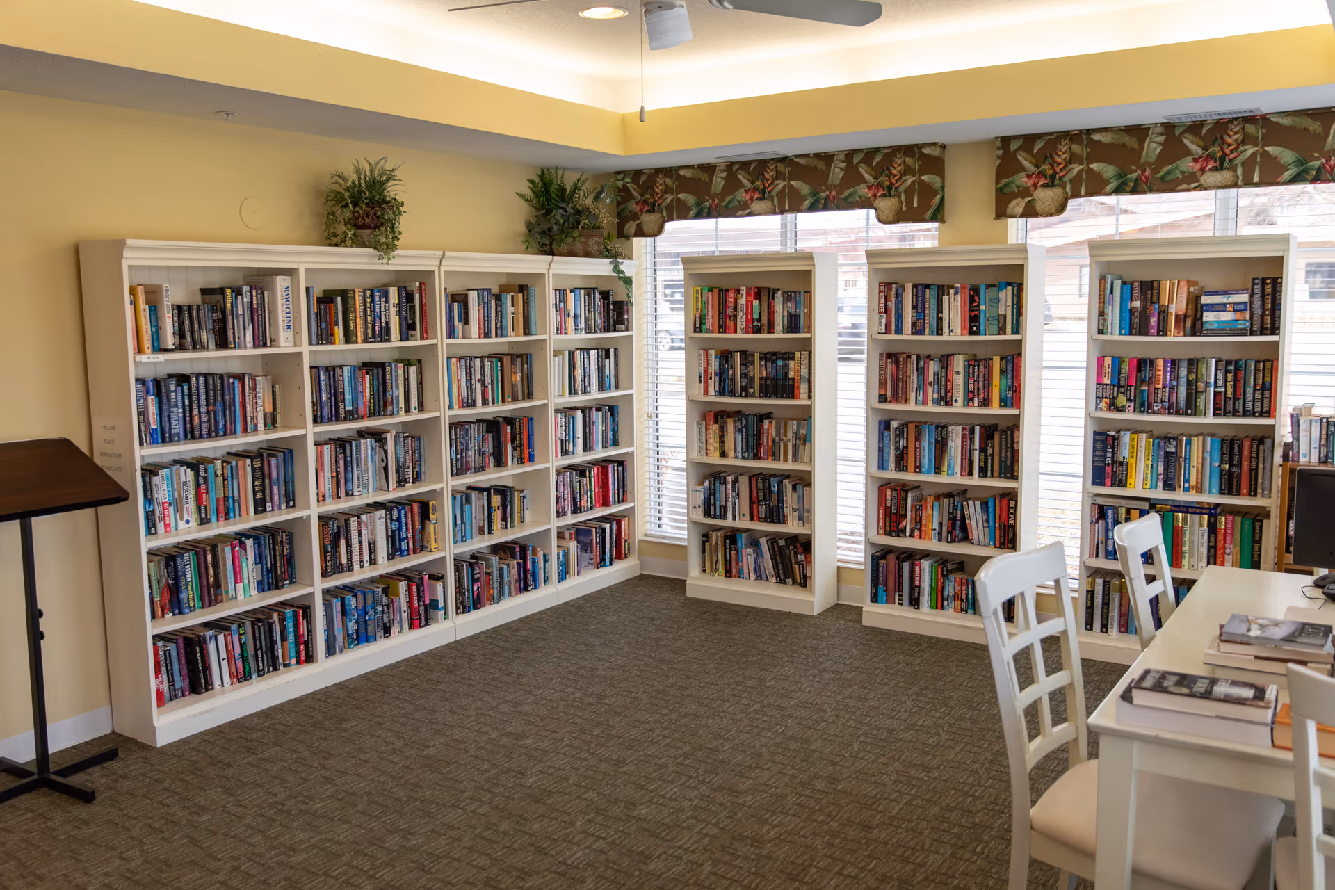 A well-lit room with multiple white bookshelves filled with books along the walls. There are windows with floral valances above the shelves, and a white table with chairs is partially visible on the right side. The room has a carpeted floor and a ceiling fan with a light fixture.