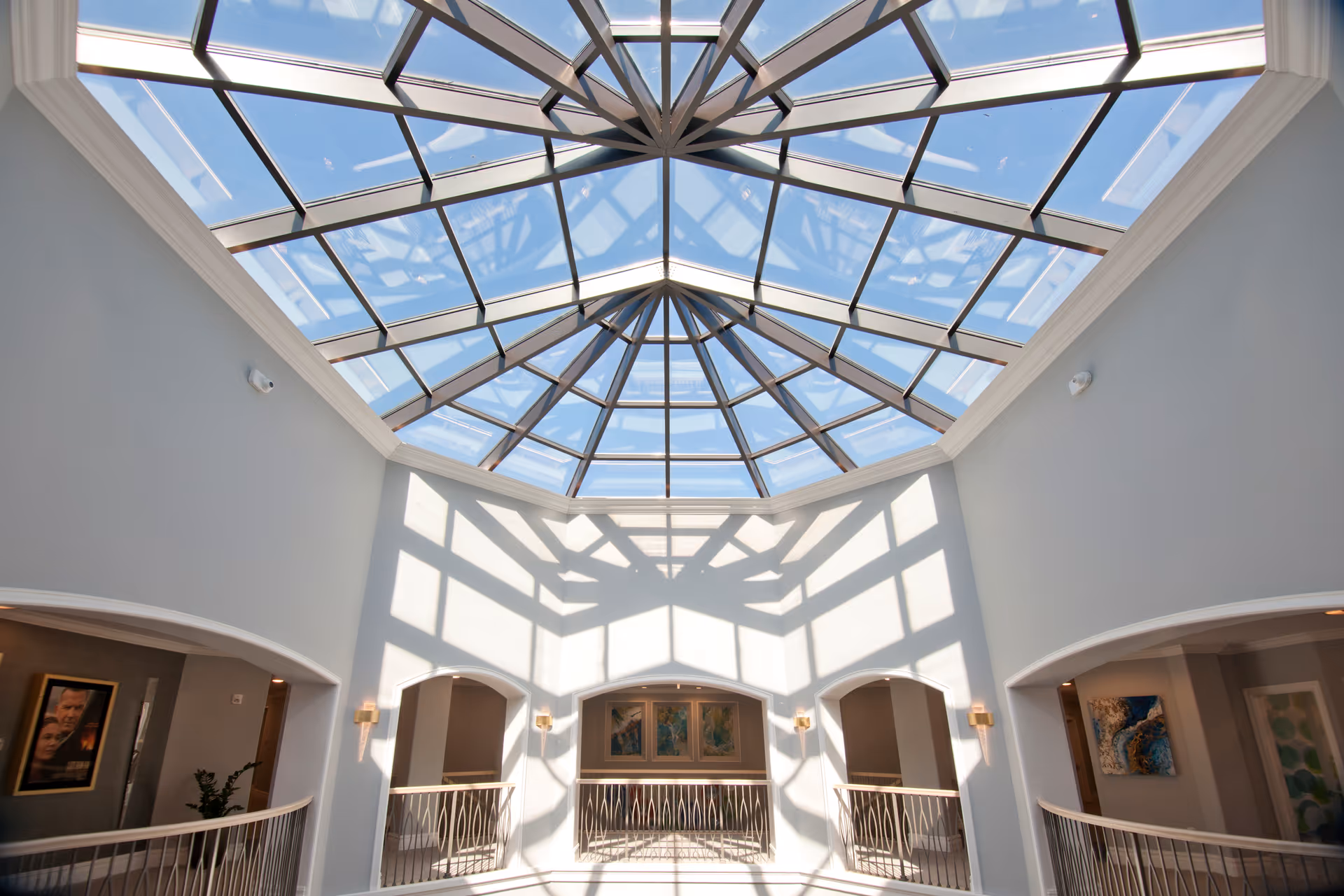 Interior view of a senior living facility featuring a large glass skylight ceiling casting geometric shadows on the white walls. The space includes a balcony with decorative railings and arched doorways leading to other rooms, with framed artwork visible on the walls.