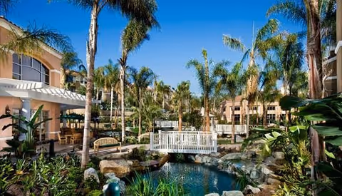 Landscaped courtyard with a pond, rocks, a white footbridge, palm trees and benches surrounded by residential buildings.