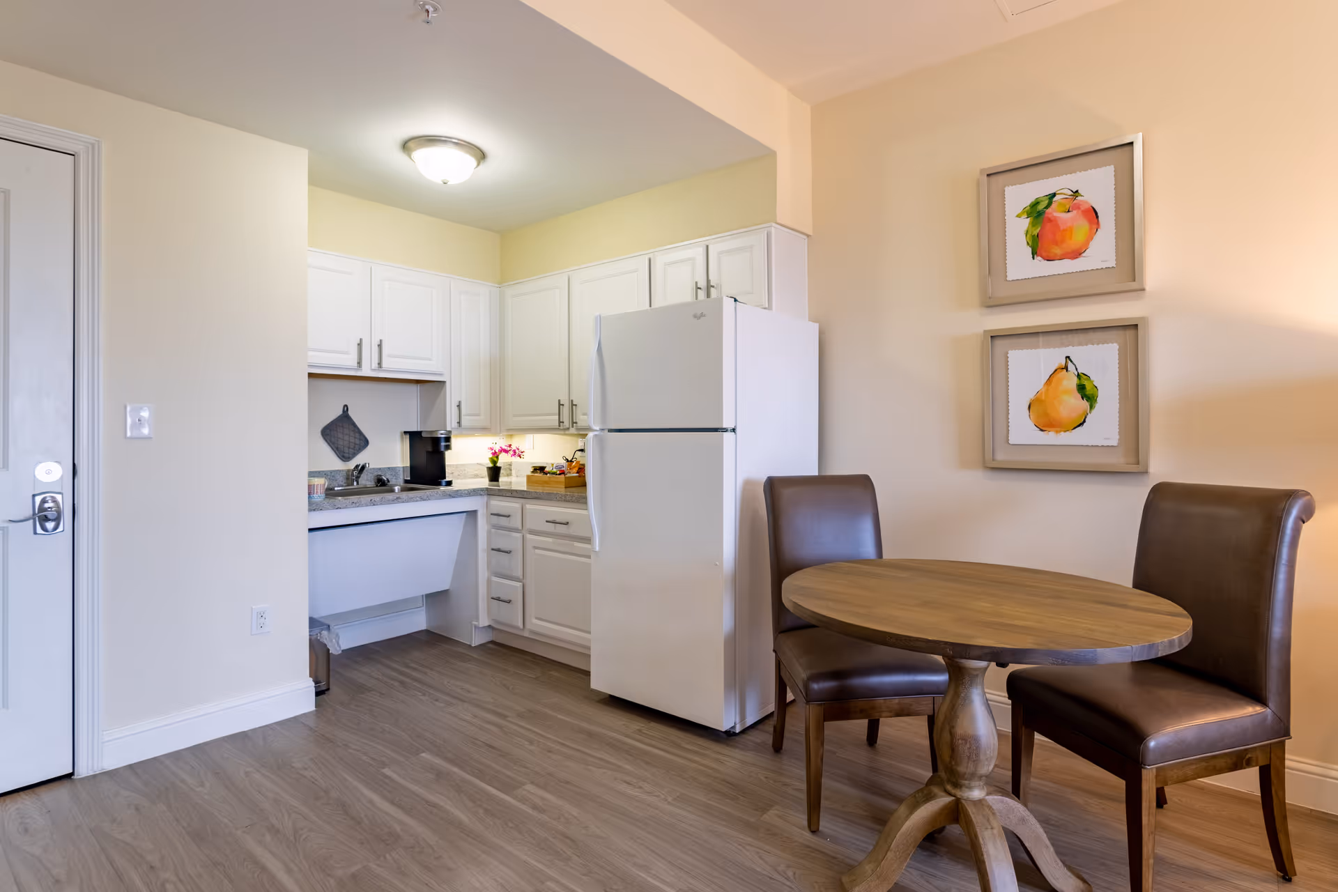 A small kitchen area with white cabinets, a white refrigerator, and a countertop with a coffee maker and a small plant. Adjacent to the kitchen is a round wooden table with two brown leather chairs. Two framed fruit paintings hang on the wall above the chairs. The floor is wood, and the walls are painted light beige.