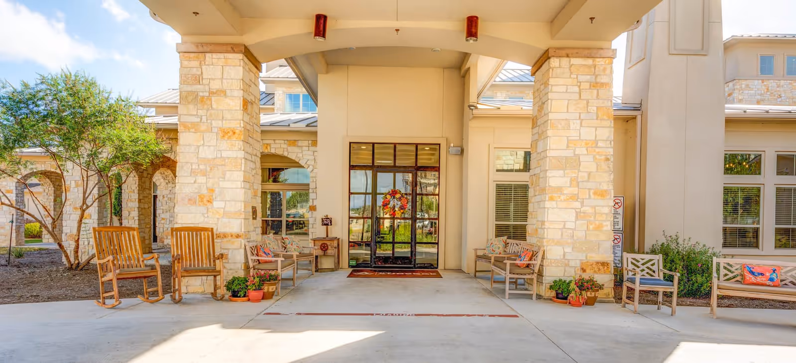 Entrance area of a senior living facility with stone pillars, a glass door decorated with a wreath, wooden rocking chairs, benches with cushions, potted plants, and a tree on the left side.