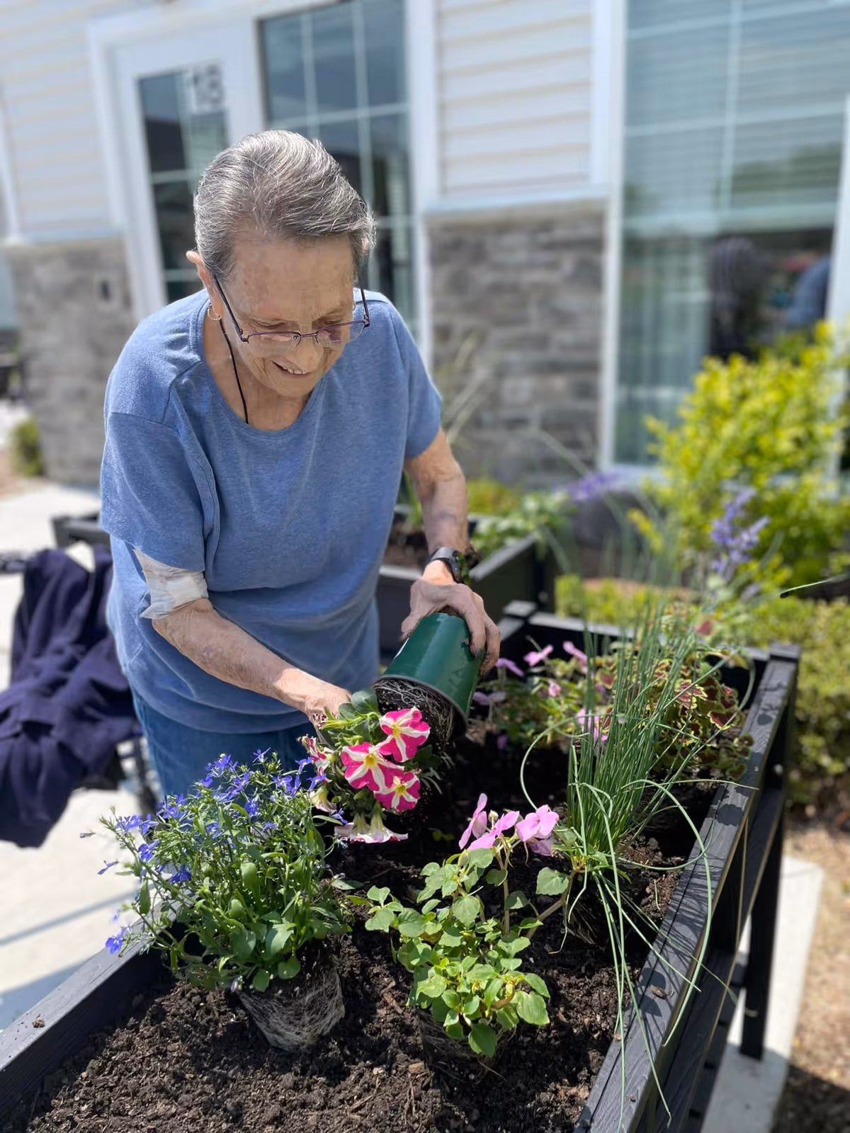 An elderly woman wearing glasses and a blue shirt is planting flowers in a raised garden bed outside a building with stone and siding exterior. She is smiling and appears to be enjoying the gardening activity on a sunny day.