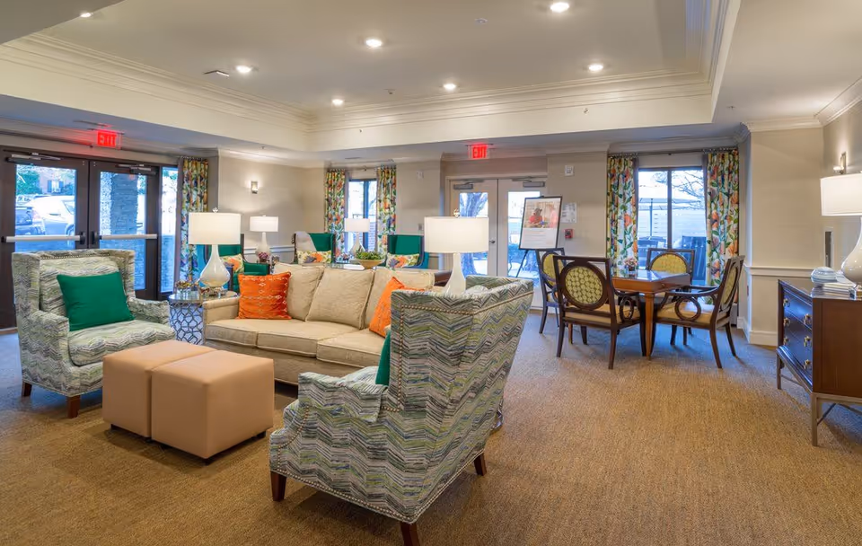 A bright and spacious living room area in a senior living facility with comfortable seating including a beige sofa with orange and green pillows, two patterned armchairs with green pillows, two beige ottomans, and several table lamps. There are large windows with floral curtains, a wooden dining table with chairs, and double glass doors leading outside.