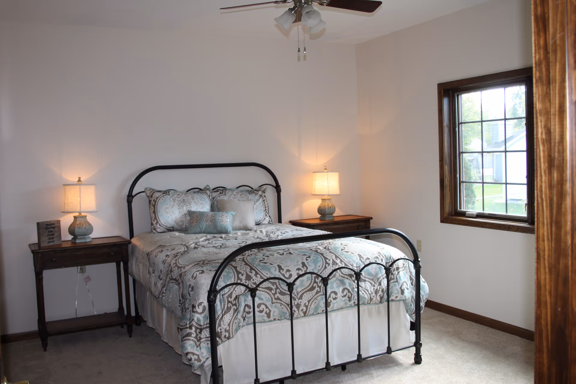 Light-filled bedroom with a metal-framed bed, patterned bedding, two bedside tables with lamps, and a window.