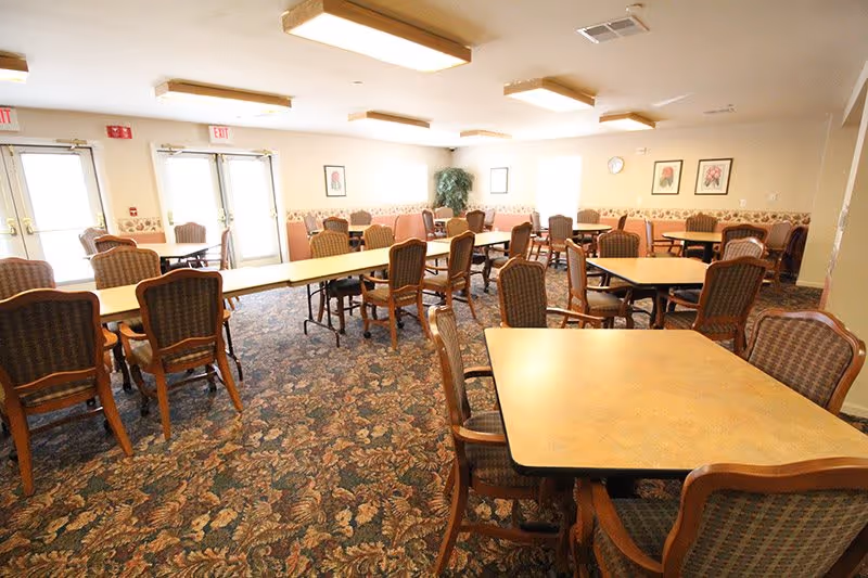 A spacious dining room with multiple rectangular and square tables surrounded by cushioned wooden chairs. The room has patterned carpet flooring, beige walls with floral wallpaper border, framed floral artwork, and large windows letting in natural light. There are double glass doors at the far end of the room.