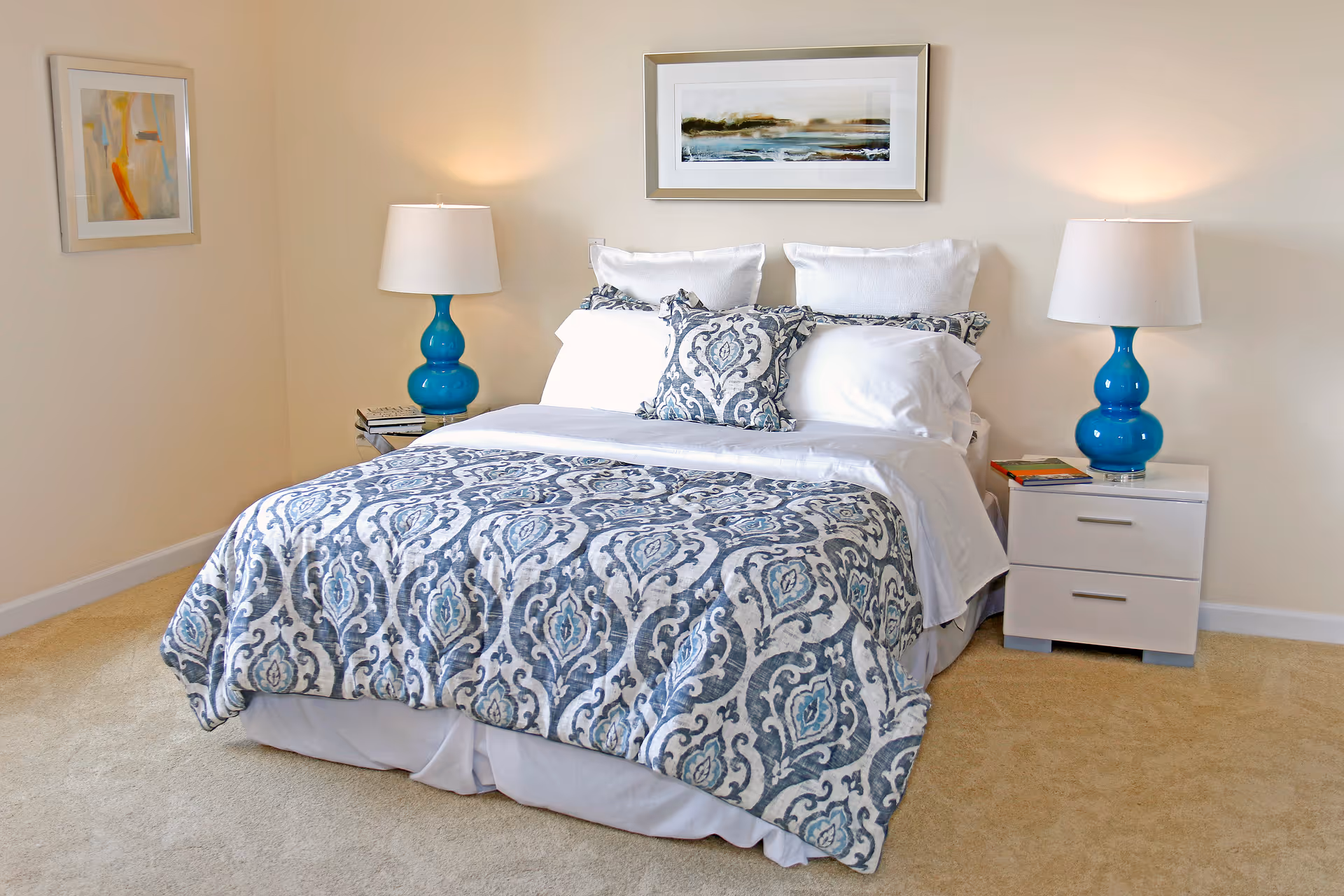 A neatly made bed with white sheets and a blue and white patterned comforter and pillows. There are two matching blue lamps on white nightstands on either side of the bed. The room has beige walls and carpet, with framed artwork hanging above the bed and on the left wall.