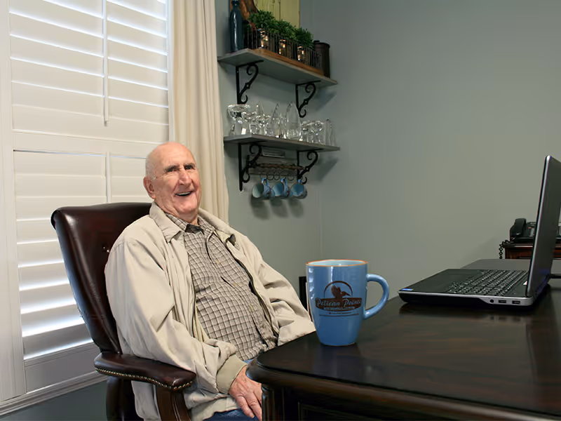 An elderly man sitting in a leather chair at a wooden desk with a laptop and a blue mug that has the Pelican Pointe logo. Behind him is a window with white shutters and shelves on the wall holding glassware and small plants.