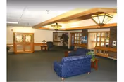 A spacious interior common area with blue upholstered armchairs, wooden furniture, and decorative ceiling lights. The room has carpeted flooring, large windows, and a wooden beam ceiling feature.
