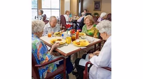 A group of elderly residents seated around a table eating meals in a communal dining room.