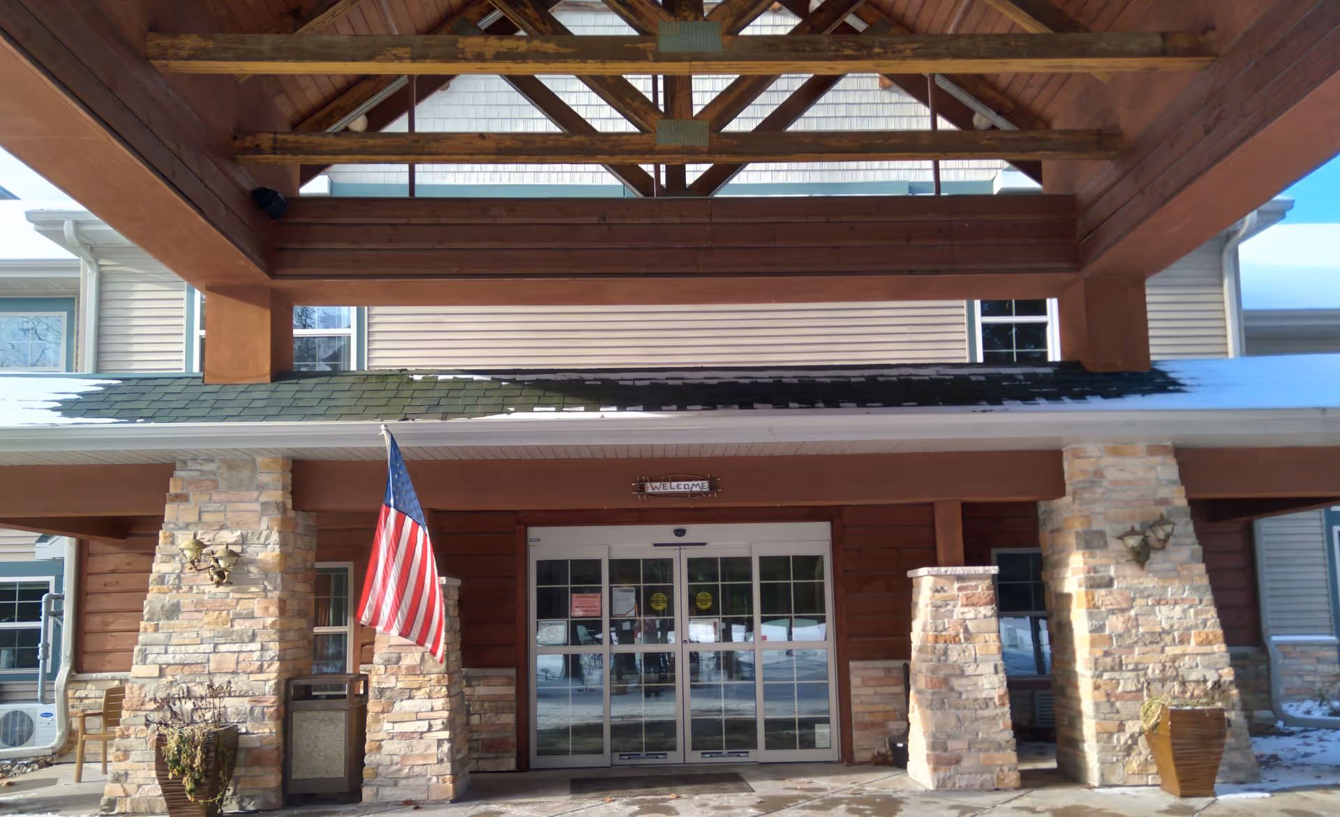 Entrance of a senior living facility with a covered wooden porch supported by stone pillars. An American flag is displayed on the left side near the entrance. The building has beige siding and large glass double doors with a small 'Welcome' sign above them. Snow is visible on the roof and ground.