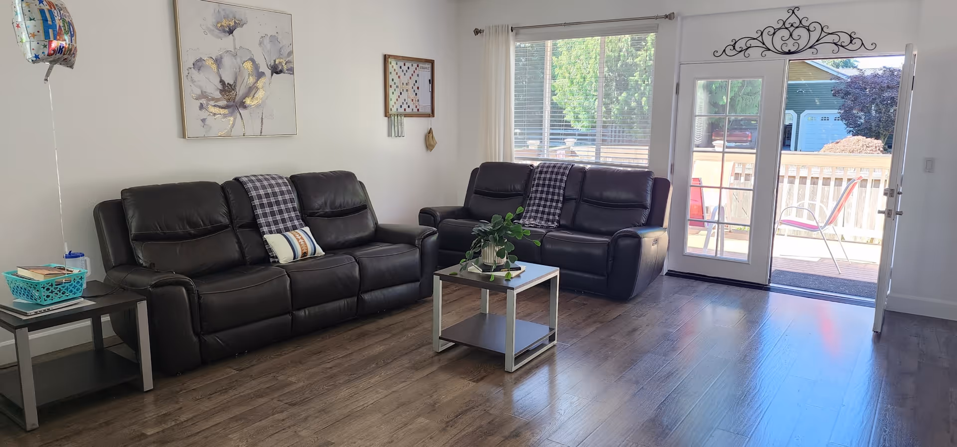 A bright living room with two black leather sofas, each with a plaid throw blanket. A small coffee table with a plant is centered between the sofas. There is a side table with books and a balloon on the left. Large window with white curtains and a glass door open to an outdoor patio with chairs and a wooden fence.
