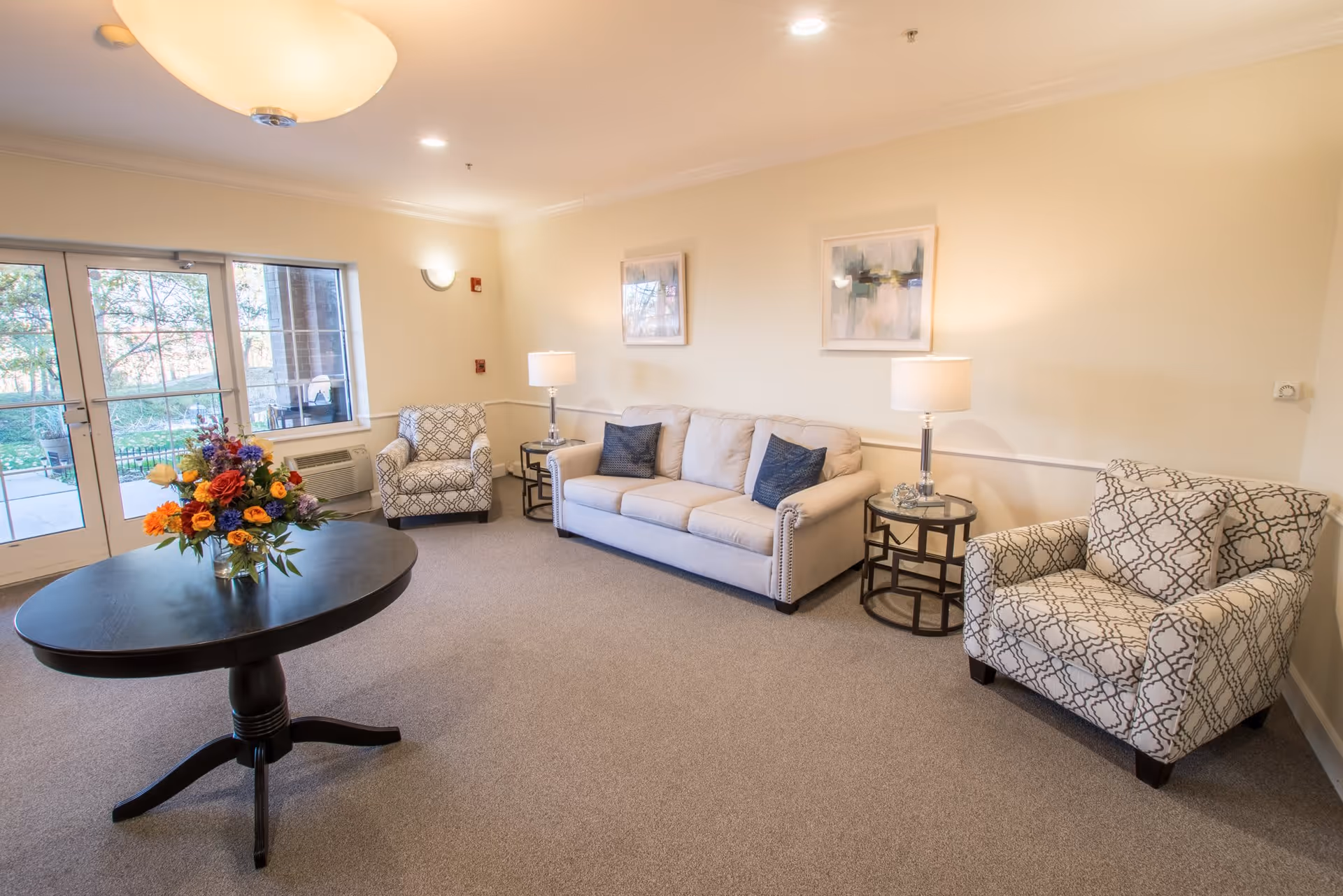 A bright and cozy living room with a beige sofa adorned with two dark blue pillows, two patterned armchairs, two side tables with lamps, a round black table with a colorful flower arrangement, and large windows and glass doors letting in natural light.