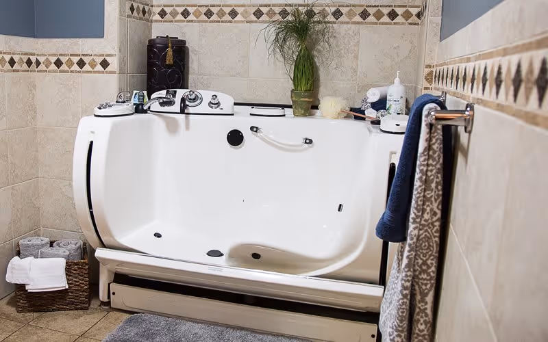 A walk-in bathtub in a tiled bathroom with beige and brown diamond-patterned accents. The tub has a built-in seat, grab bars, and controls on the rim. Nearby are a basket with rolled towels, a blue towel hanging on a rack, and various bath items including a plant and soap dispenser on the tub edge.