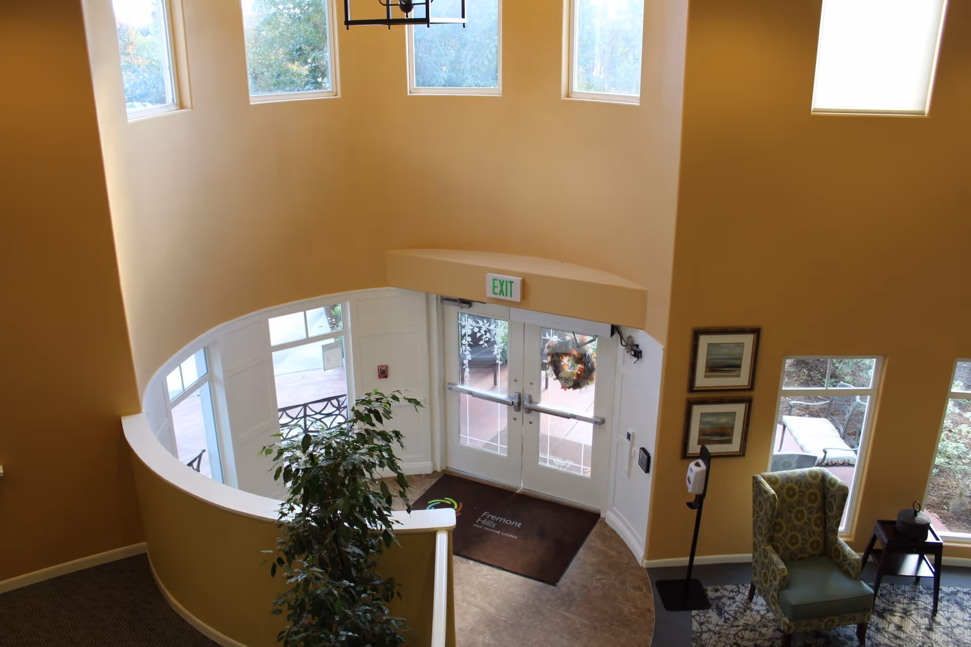 Interior view of a senior living lobby from above, featuring a curved balcony, potted plant, double glass exit doors with a wreath, and a seating area.