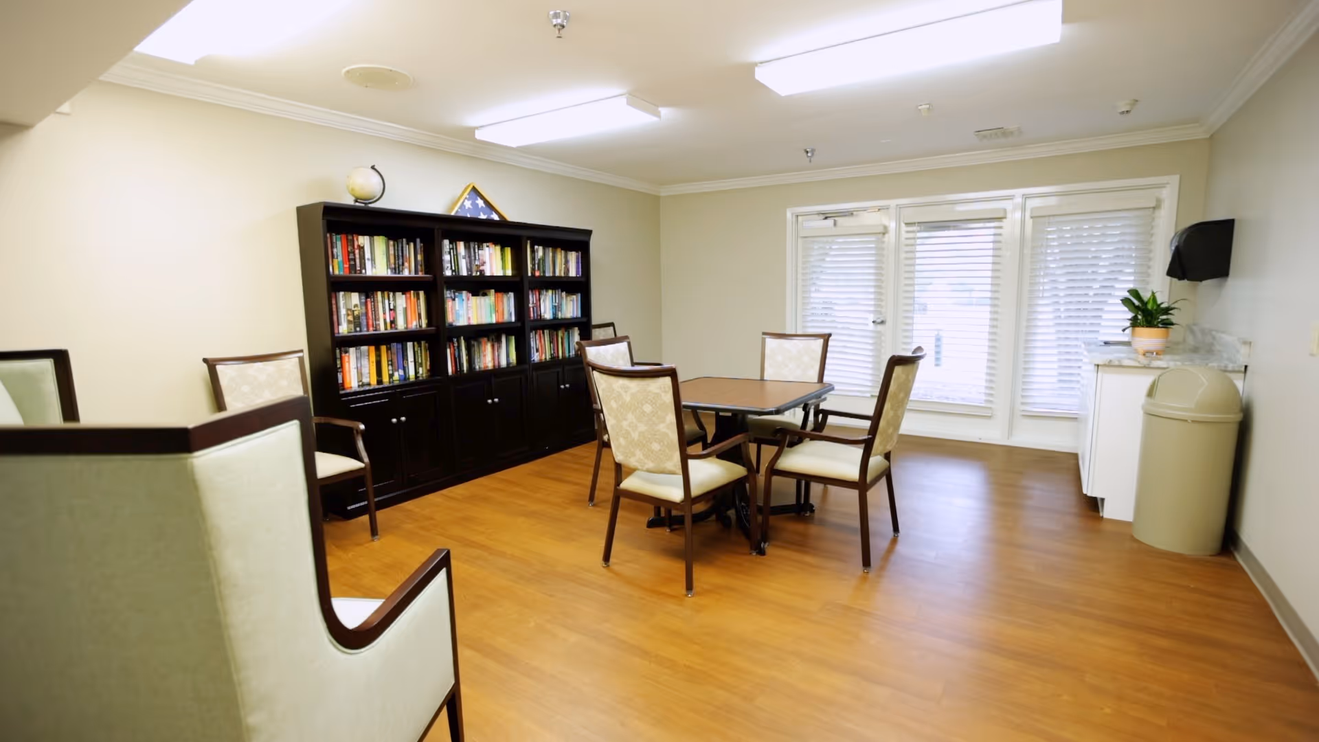 Bright common room with a table and chairs, a bookshelf filled with books, and large windows/doors.