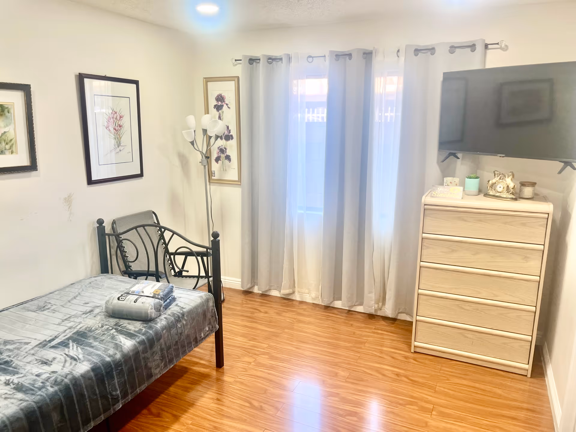 A small bedroom with a single bed covered in a gray blanket, a black metal chair next to the bed, and a wooden dresser with four drawers. A flat-screen TV is mounted on the wall above the dresser. The room has wooden flooring, white walls with framed floral artwork, and a window with sheer white curtains allowing natural light to enter.