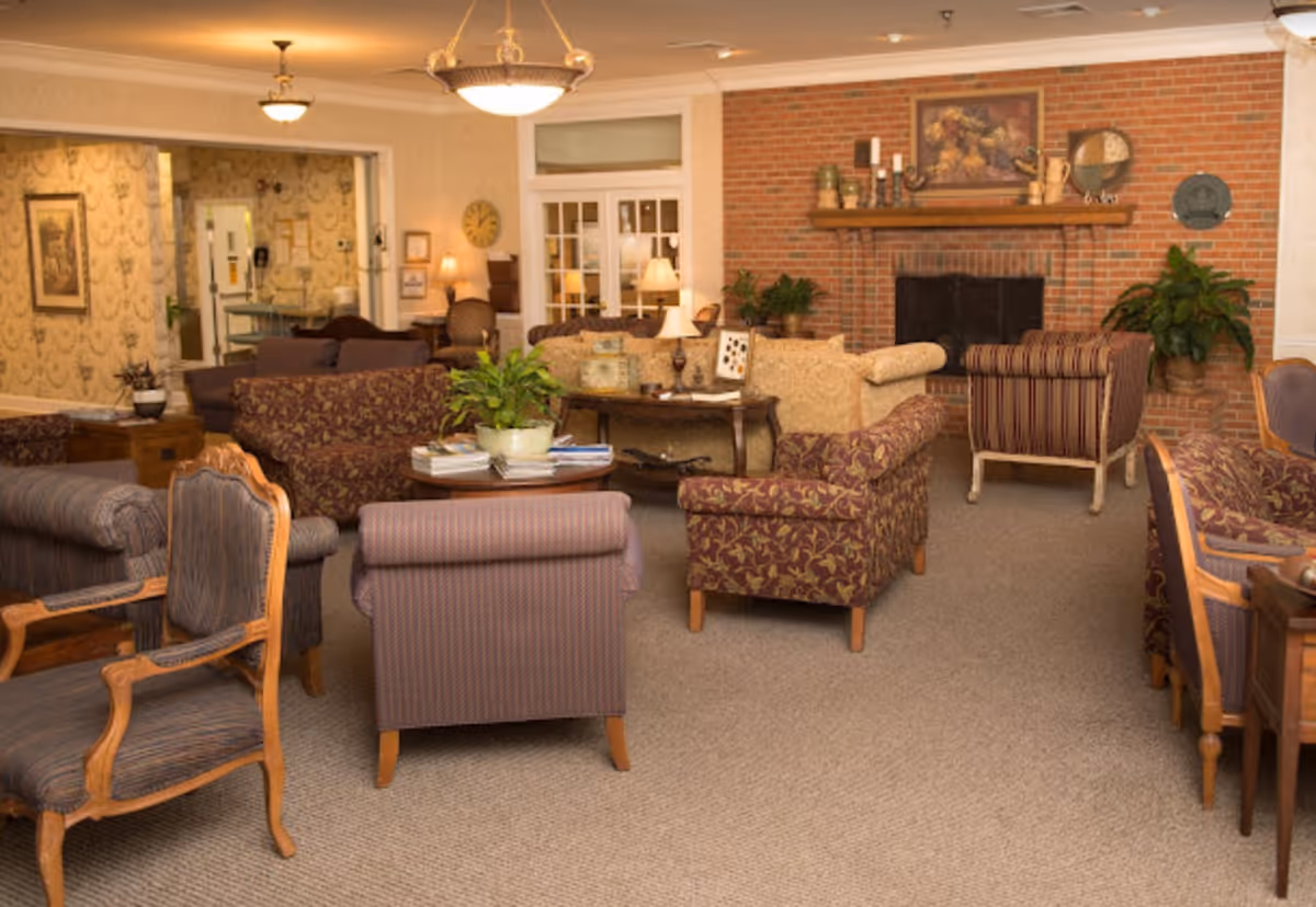 A cozy senior living facility common area with multiple upholstered armchairs and sofas arranged around a brick fireplace. The room is warmly lit with ceiling lights and table lamps, decorated with plants, framed artwork, and a clock on the wall. The carpeted floor and traditional furniture create a comfortable and inviting atmosphere.