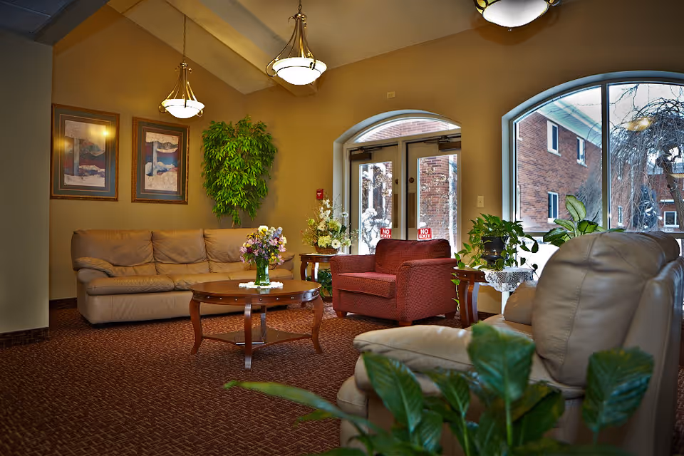 A cozy seating area in a senior living facility with a beige leather sofa, a red armchair, and a beige recliner. There is a wooden coffee table with a vase of flowers in the center, and side tables with plants and floral arrangements. The room has warm lighting from ceiling fixtures, framed artwork on the walls, and large windows showing an exterior brick building and trees.
