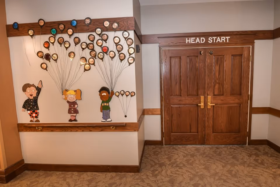 Hallway interior showing double wooden doors labeled "HEAD START" and a wall mural of cartoon children with balloon decorations.