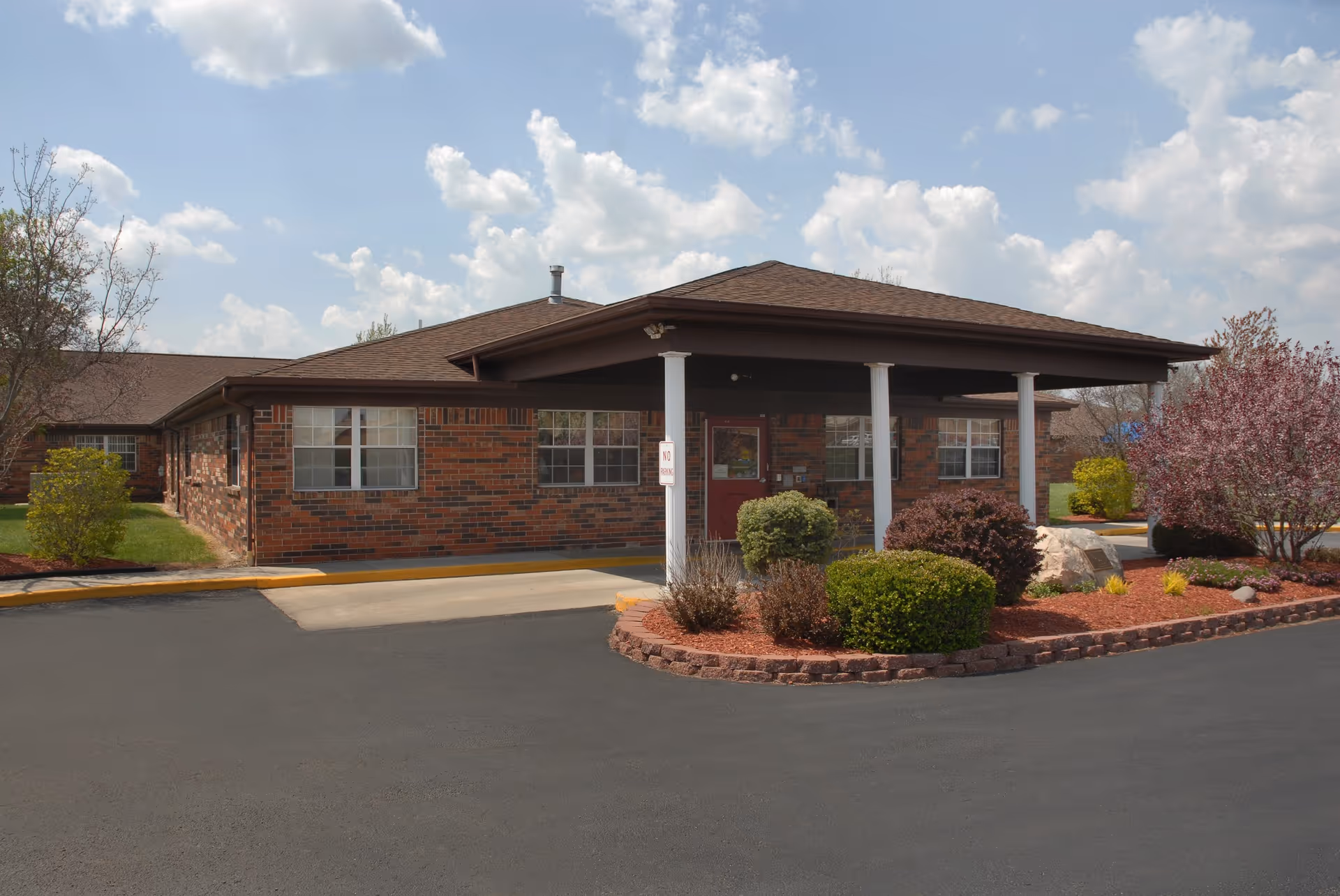Single-story brick building front entrance with a covered drive, landscaped beds, and a paved driveway under a partly cloudy sky.