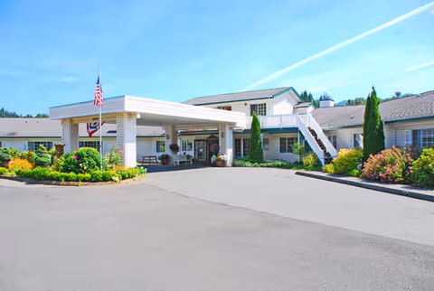 Exterior view of Brookdale Fairhaven senior living facility showing a single-story building with a covered entrance, an American flag, landscaped bushes and flowers, and a clear blue sky.