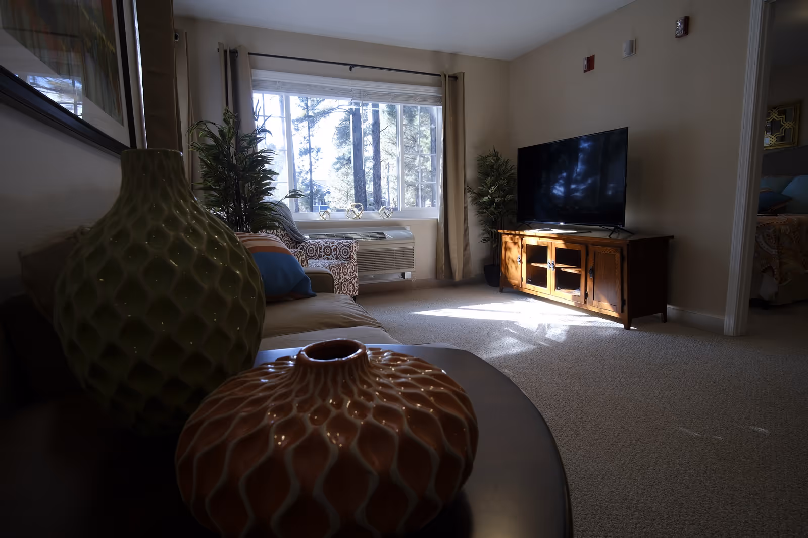 Living room with decorative vases in the foreground, a sofa, a TV on a wooden stand, and a large window showing trees.